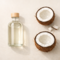 Clear glass bottle of coconut oil with fresh coconut halves in a minimal natural skincare flat-lay under soft daylight