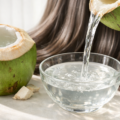 Fresh green coconut cut open with clear coconut water being poured into a glass bowl, natural shiny hair texture in the background under soft daylight.