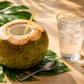 Fresh coconut with straw and glass of cold water, tropical leaves, small spoon on light wooden table, warm morning sunlight